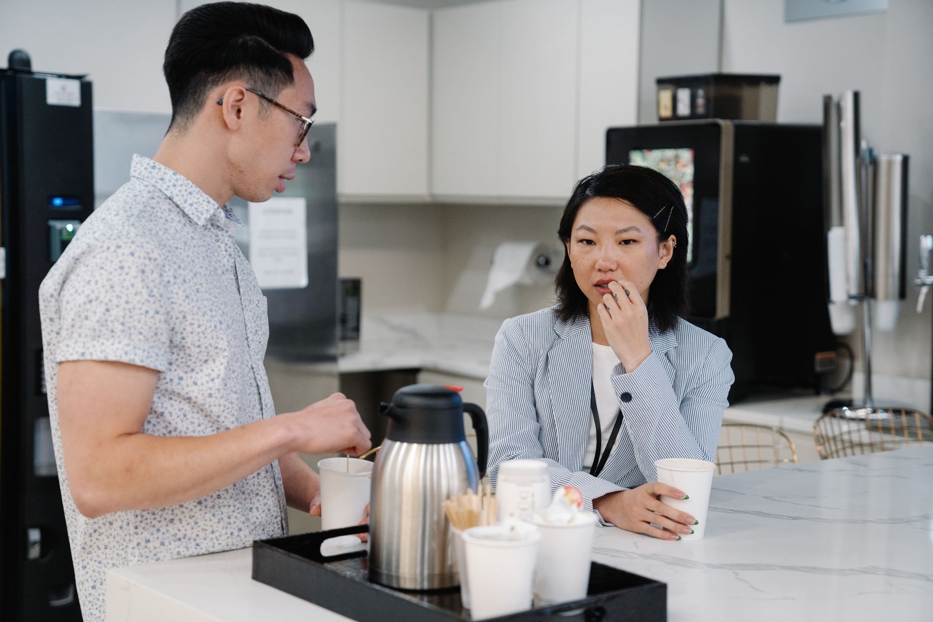 Two colleagues discussing over coffee in a modern office break room setting.