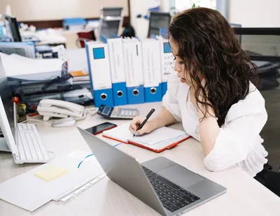 An office worker takes notes while working at a desk with a laptop and documents.